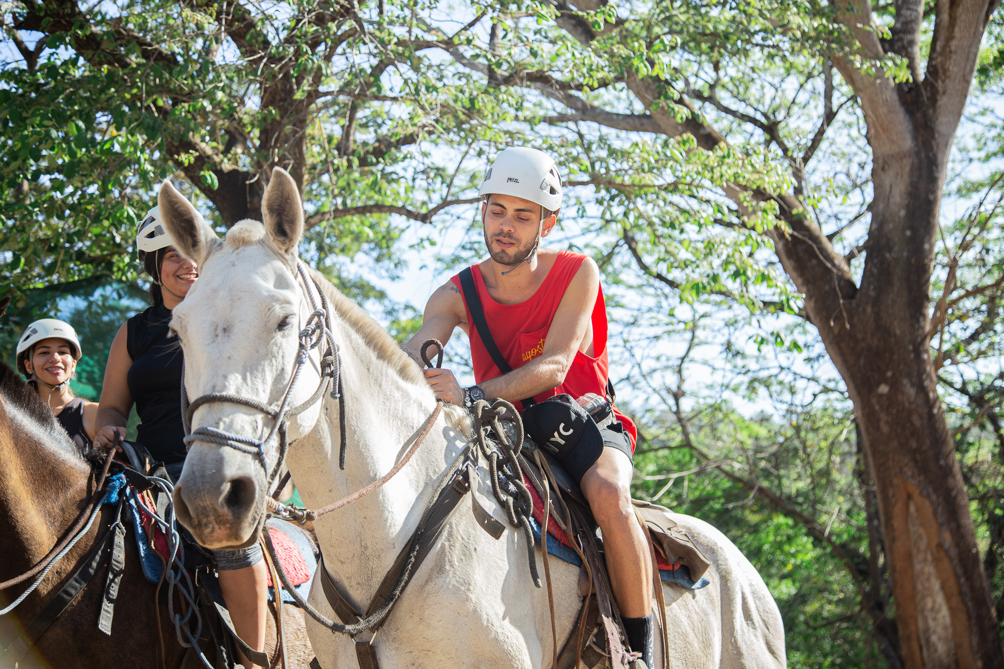 People riding horses in a wooded area, wearing helmets and casual attire.