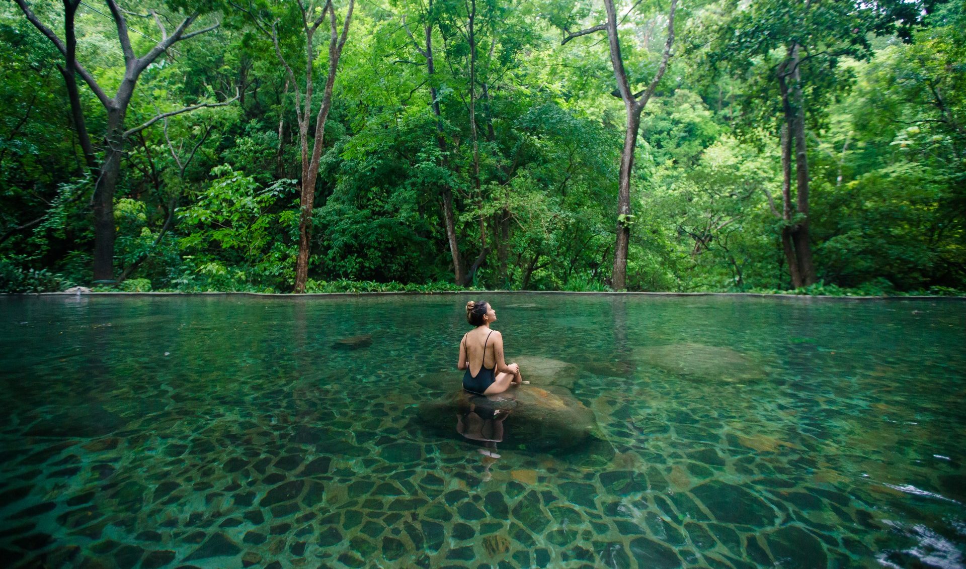 Person sits on rock in clear, forest-surrounded stream, facing lush greenery