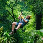 Person zip-lining through lush green forest, wearing a helmet and gloves.
