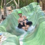 Person on a green water slide through a forest, smiling and wearing a green helmet.
