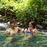 Couple in pool with drinks, surrounded by lush greenery and a flowing stream.