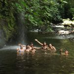 Group of people swimming in a forest stream near a small waterfall.