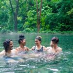 Four people swimming and laughing in a forested hot spring.