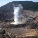 Aerial view of a smoking volcano crater surrounded by rugged terrain.