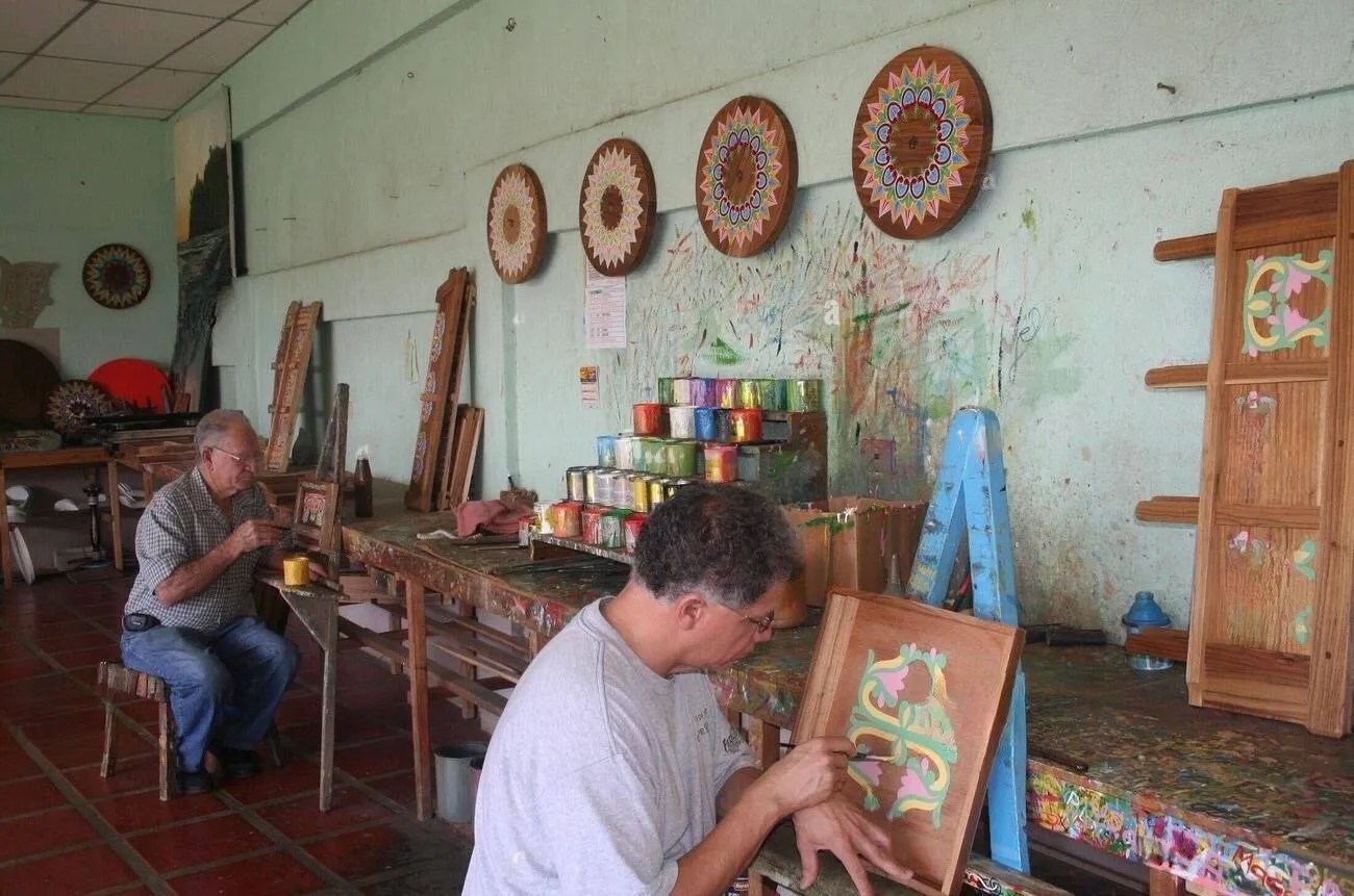 Two artisans paint wood crafts in a workshop with colorful designs on the wall.