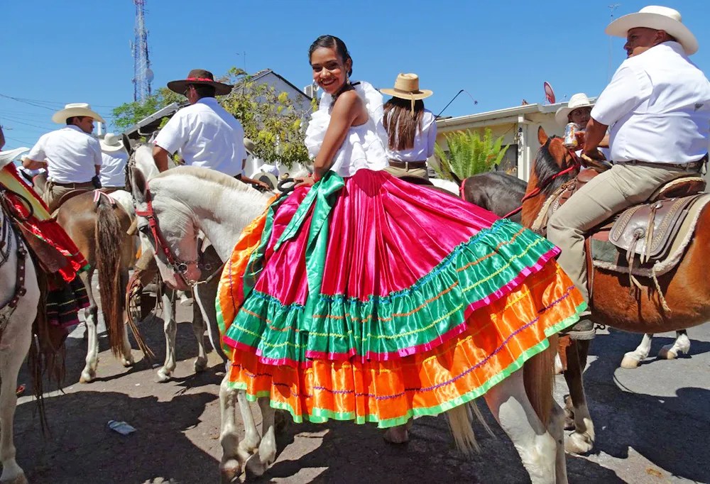 Woman in colorful dress on horseback at outdoor event with others in white shirts and hats.