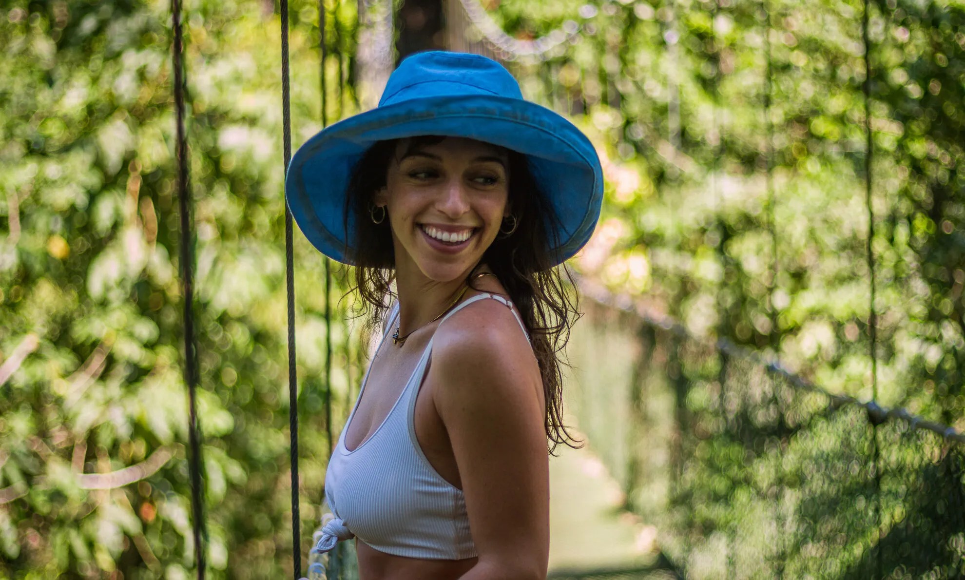 Woman in blue hat smiling on a suspension bridge with lush greenery.