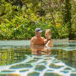 Couple in a pool surrounded by lush greenery with partial sunlight reflections on water.