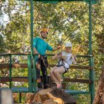 Two people in helmets and harnesses on a zipline platform in a forested area.