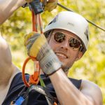 Person ziplining with helmet and sunglasses, smiling.