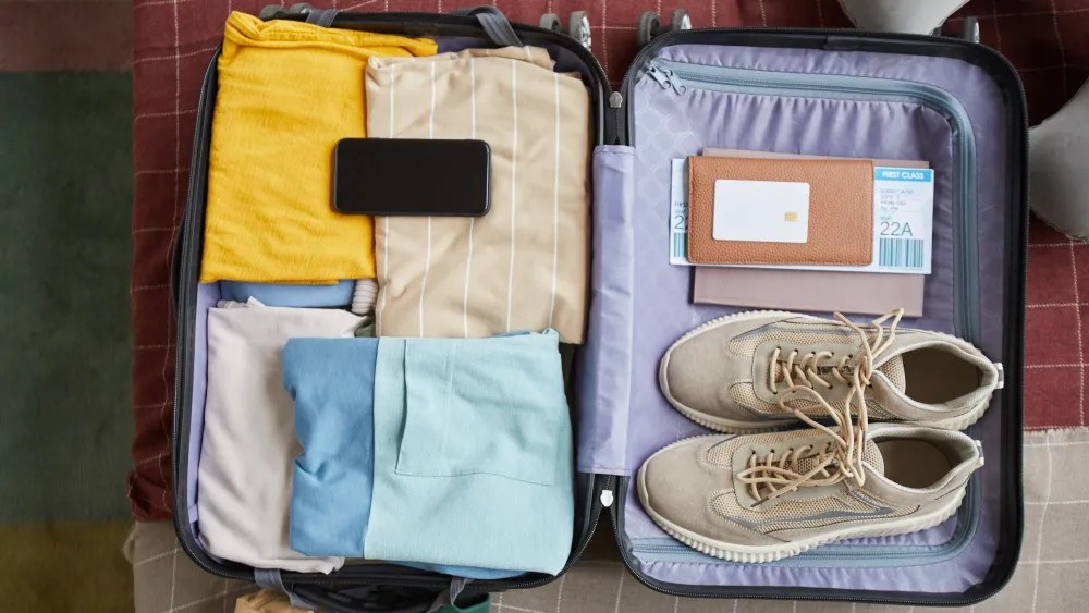 Open suitcase with clothes, shoes, phone, and travel documents on a bed.