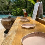 Outdoor counter with clay dishes, brush holder, menu, and pool in background under a sunny sky.