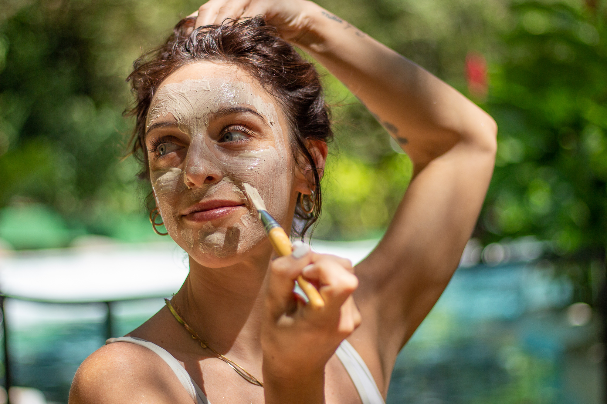 Mujer aplicándose una mascarilla facial con un pincel al aire libre en un jardín.