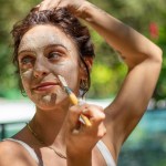 Mujer aplicándose una mascarilla facial con un pincel al aire libre en un jardín.