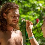 Person applying white face paint to a smiling man in a lush outdoor setting.