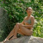 Person in a bikini sitting on a rock with lush greenery in the background.