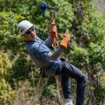 Persona haciendo tirolina en el bosque, con casco y guantes, haciendo el gesto de pulgar arriba.