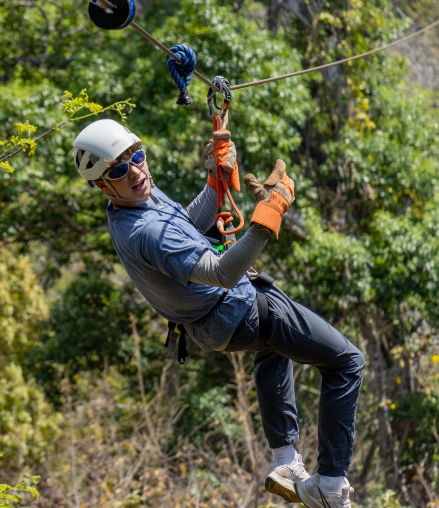 Person zip-lining in forest, wearing helmet and gloves, giving thumbs-up.