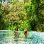 Two women in swimsuits standing in a lush, outdoor pool.