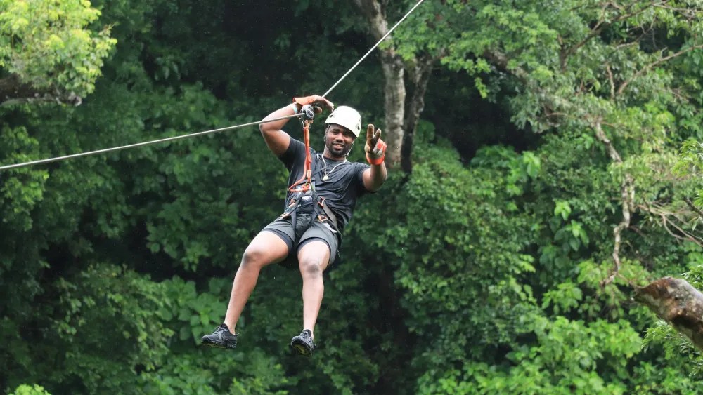 Person zip-lining through lush green forest, wearing helmet and harness.