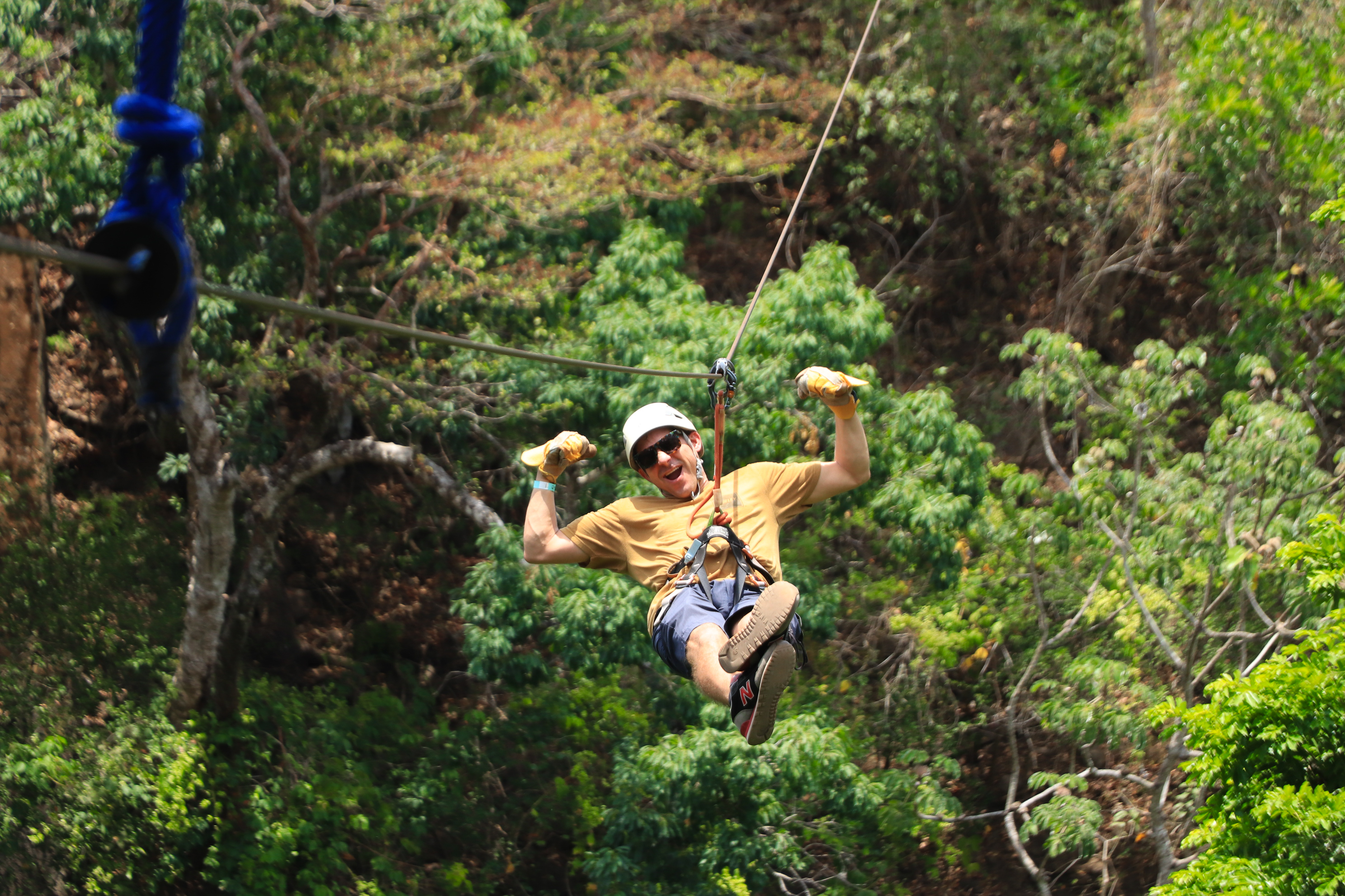Persona en tirolina por un bosque con casco y guantes, rodeada de vegetación.