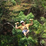 Persona en tirolina por un bosque con casco y guantes, rodeada de vegetación.