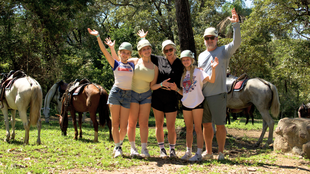 Five people posing happily in front of horses in a wooded area.