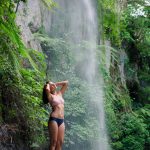 Woman in swimwear standing by a waterfall, surrounded by lush greenery.