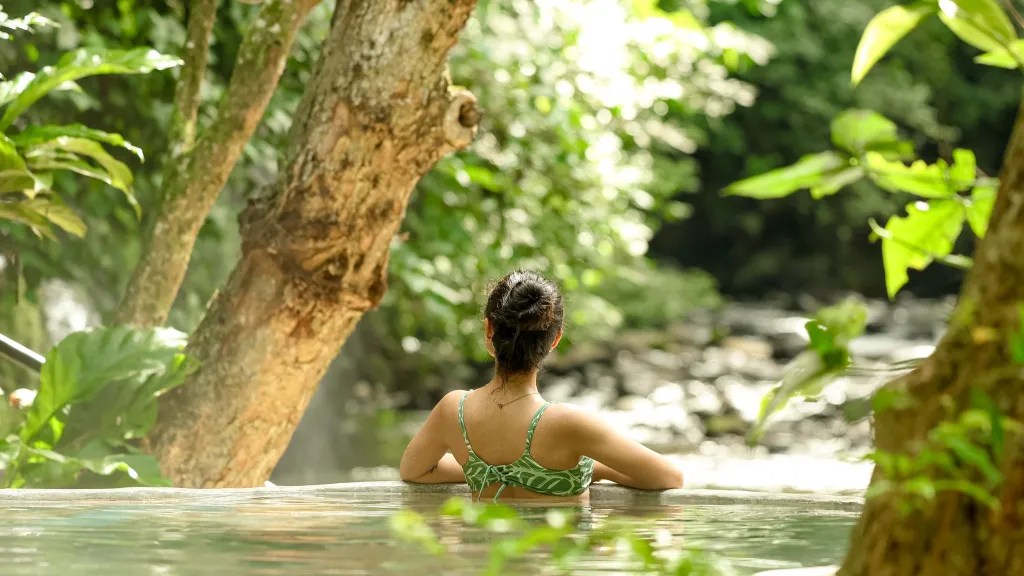 Woman in a green swimsuit relaxing in natural hot spring surrounded by lush greenery.