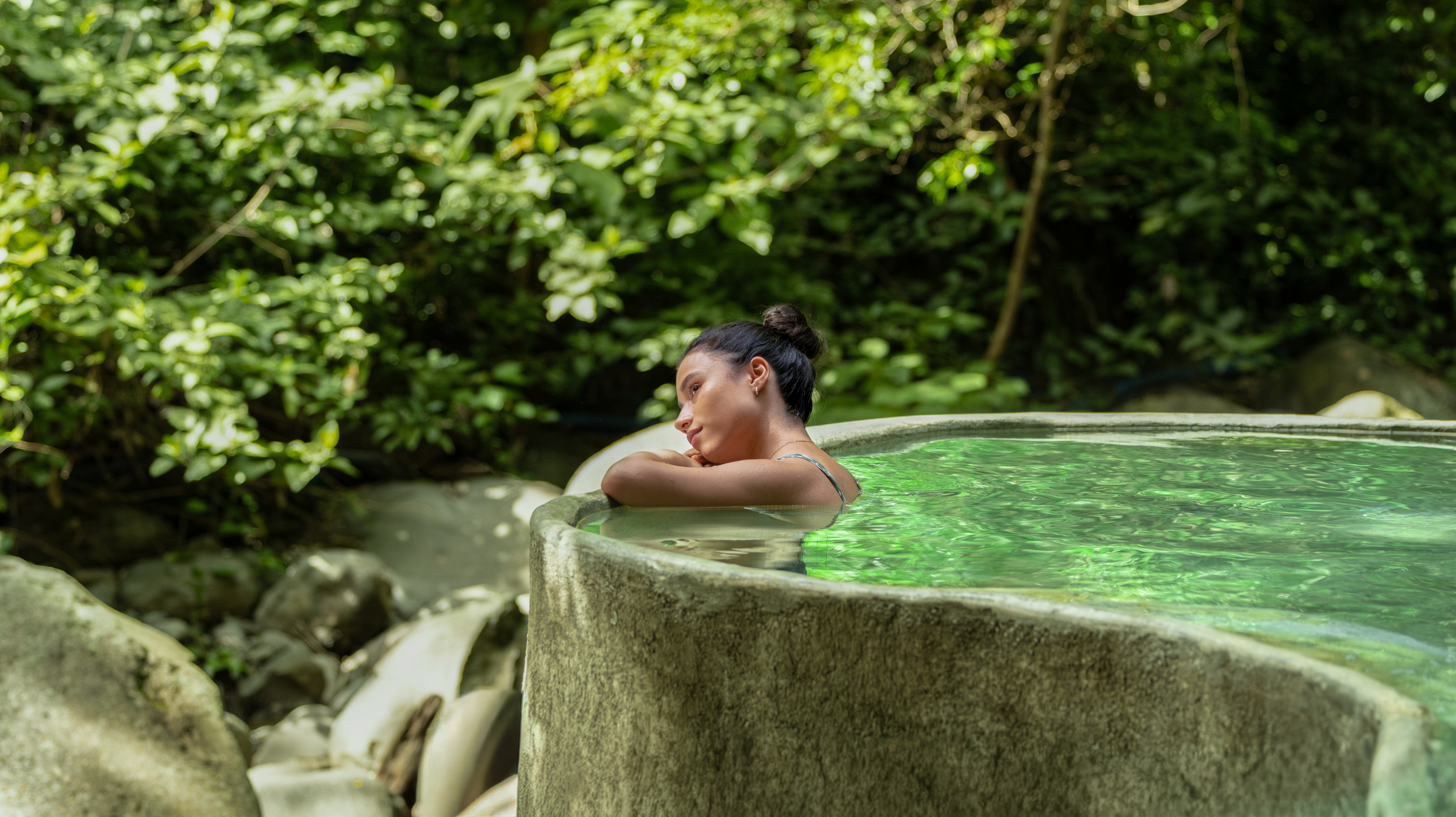 Persona relajándose en una piscina de rocas al aire libre rodeada de exuberante vegetación.