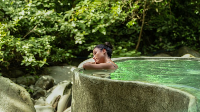 Person relaxing in an outdoor rock pool surrounded by lush greenery.