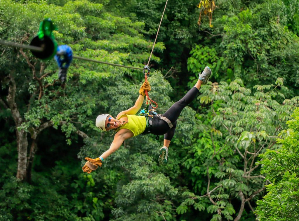 Person in yellow top zip-lining through lush green forest, wearing gloves and helmet.