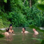 Four people swimming in a lush forest pond, surrounded by greenery.