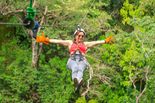 Person ziplining through forest with arms outstretched, wearing gloves and a harness.
