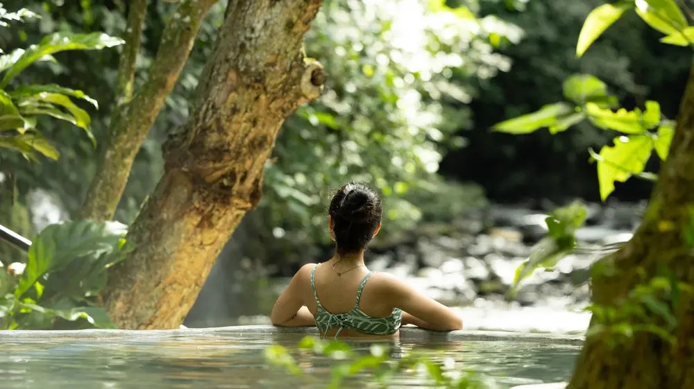 Persona en una piscina rodeada de exuberante vegetación mirando a un arroyo boscoso.