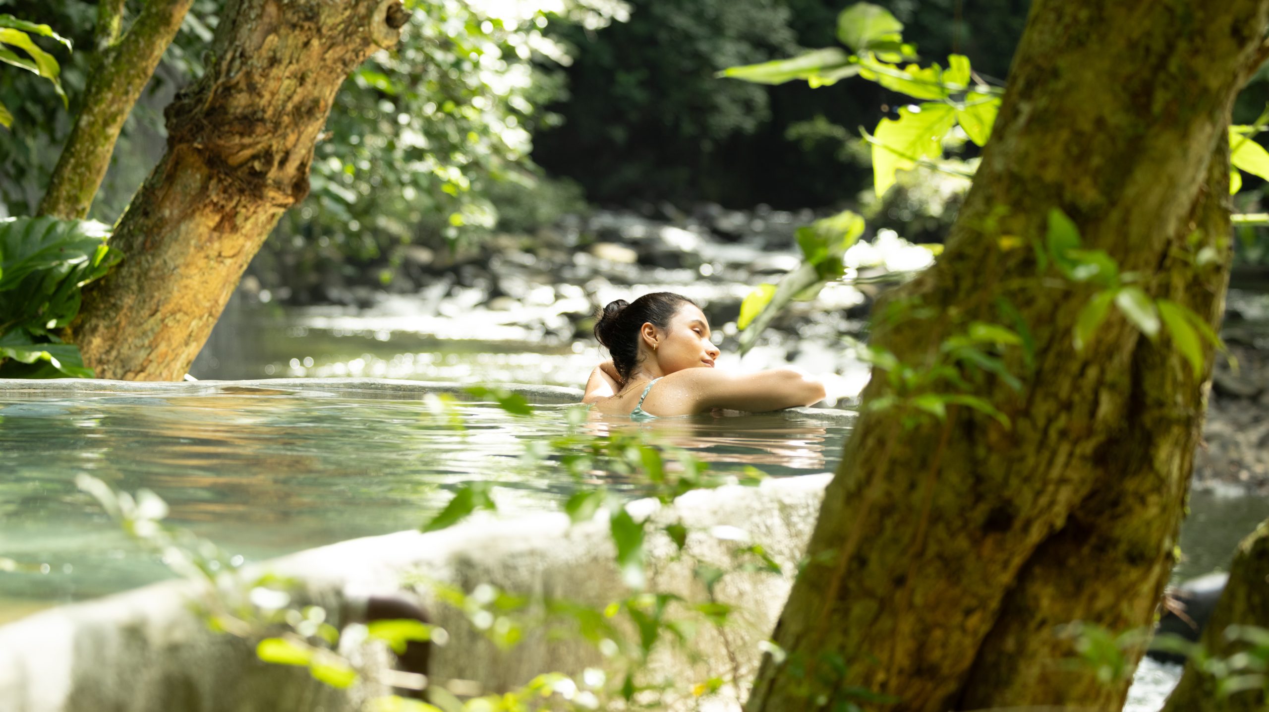 Person relaxing in an outdoor pool by a forest stream.