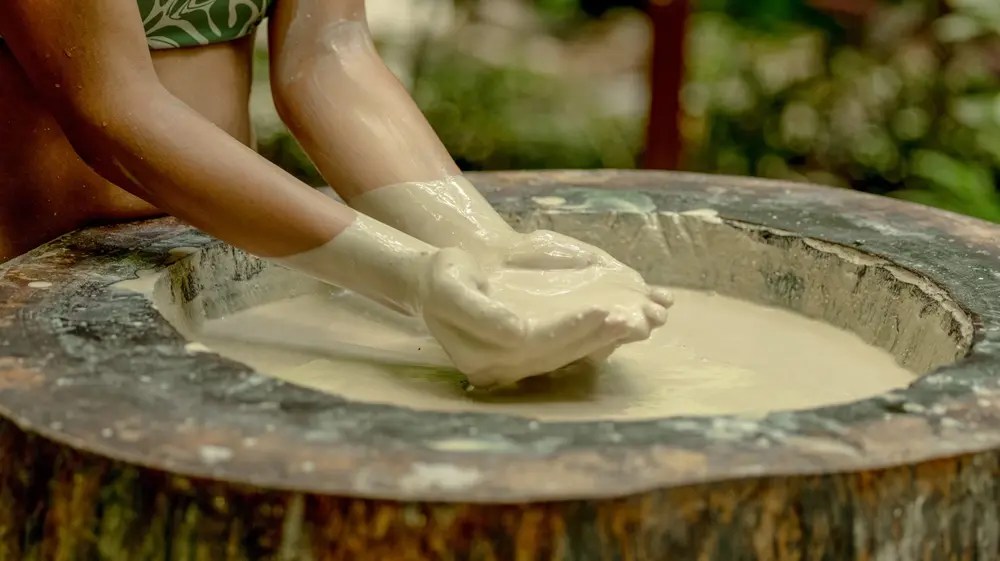 Hands stirring thick liquid in a large wooden bowl outdoors.