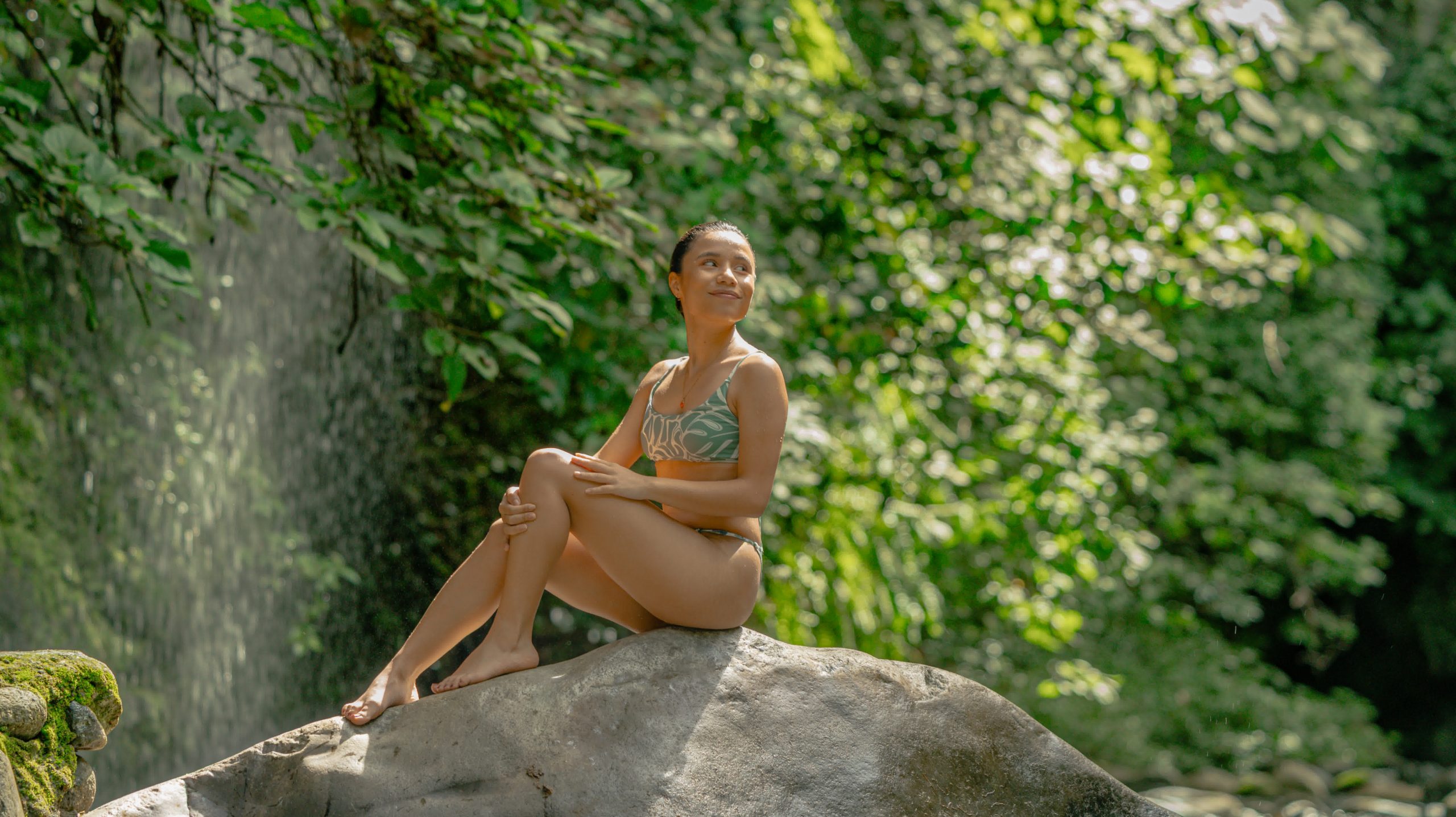 Woman in swimwear sitting on rock by waterfall in lush forest.