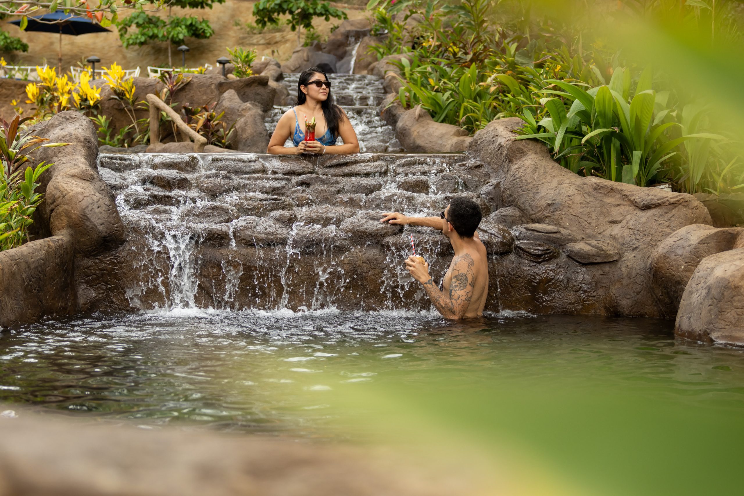 Dos personas se relajan en una piscina con cascada revestida de piedra y rodeada de exuberante vegetación.