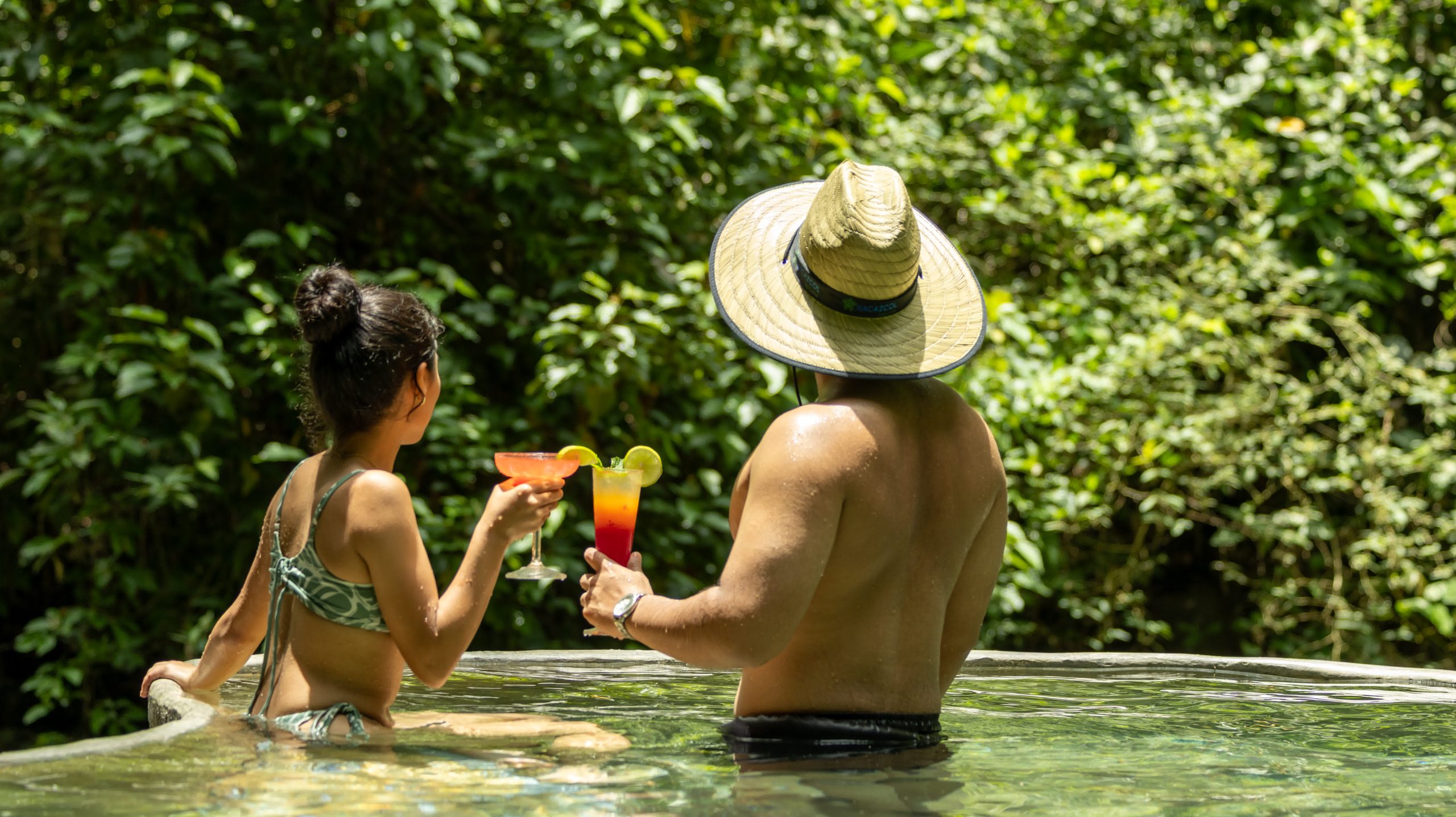 Two people enjoying tropical drinks while relaxing in a volcanic pool surrounded by lush greenery at Rincon Thermals