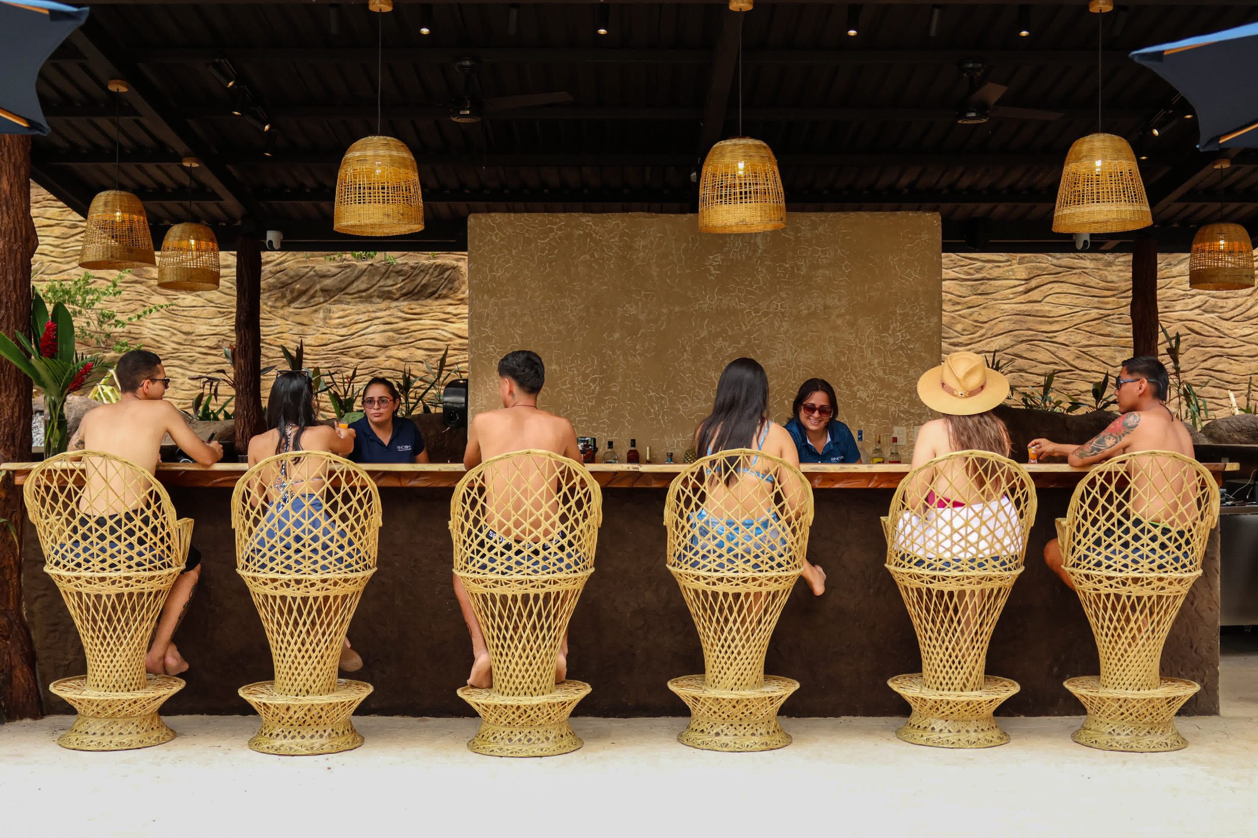 People seated at an outdoor bar with rattan chairs and hanging lights.