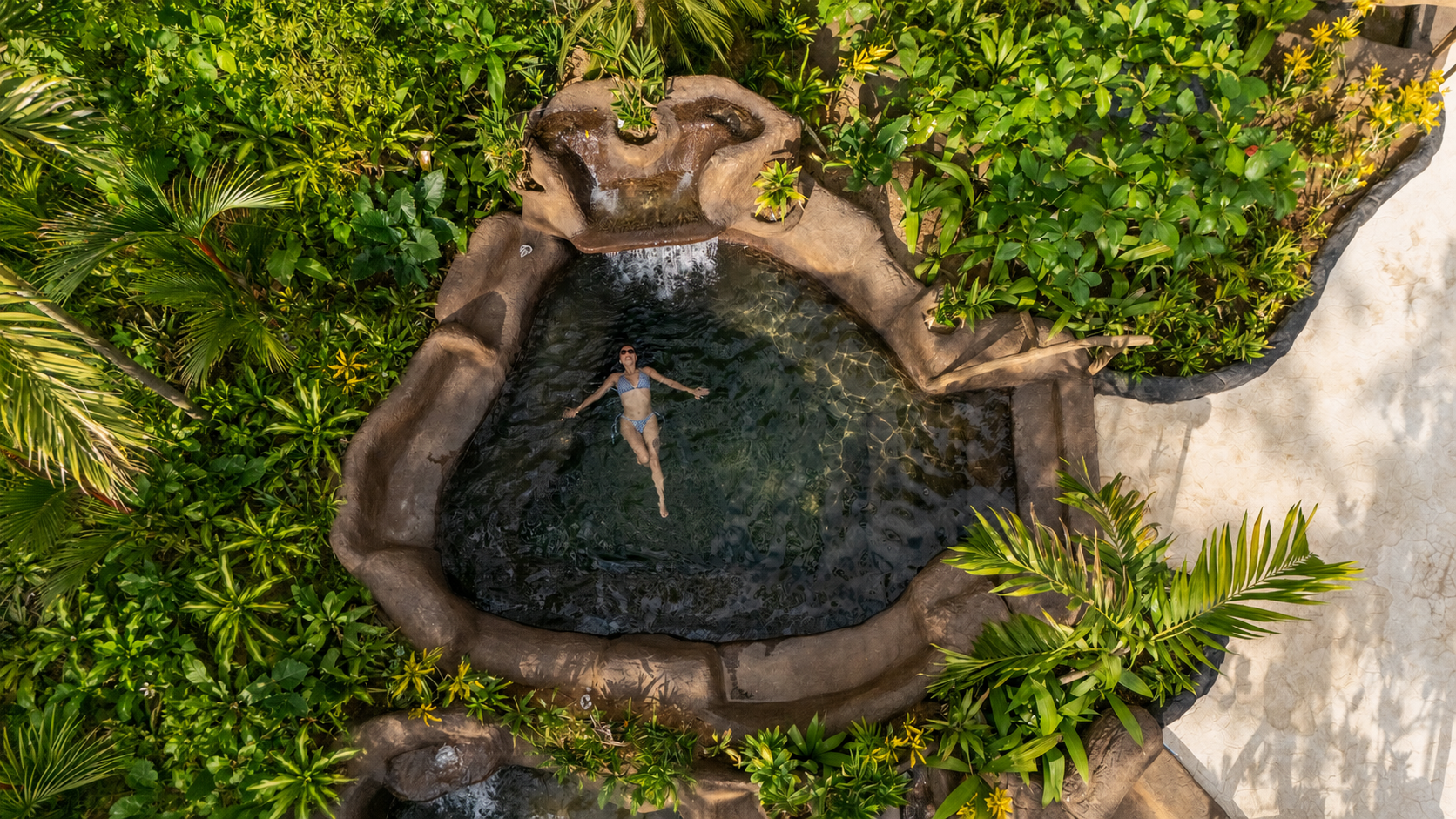 Person relaxing in a circular rock pool surrounded by lush greenery.