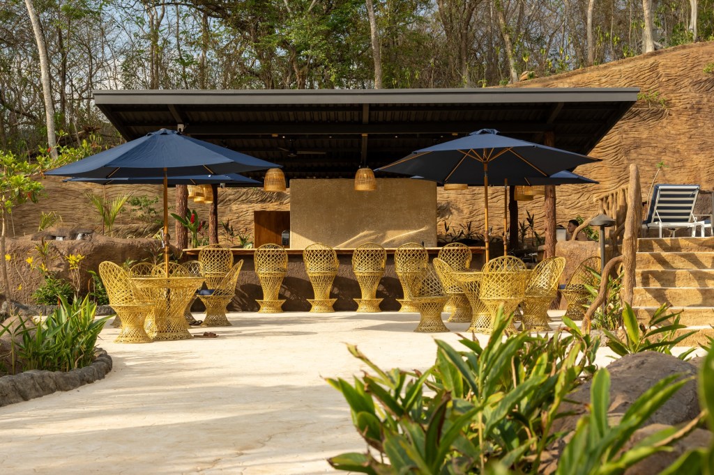 Outdoor bar with wicker chairs, blue umbrellas, and lush greenery.
