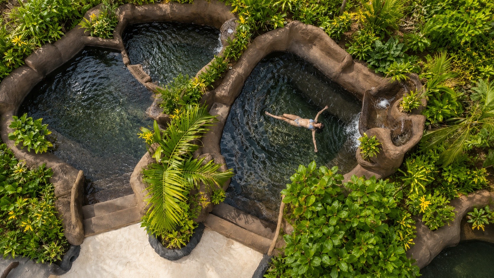 Person swimming in a series of volcanic rock-lined pools surrounded by lush greenery at Rincón Thermals