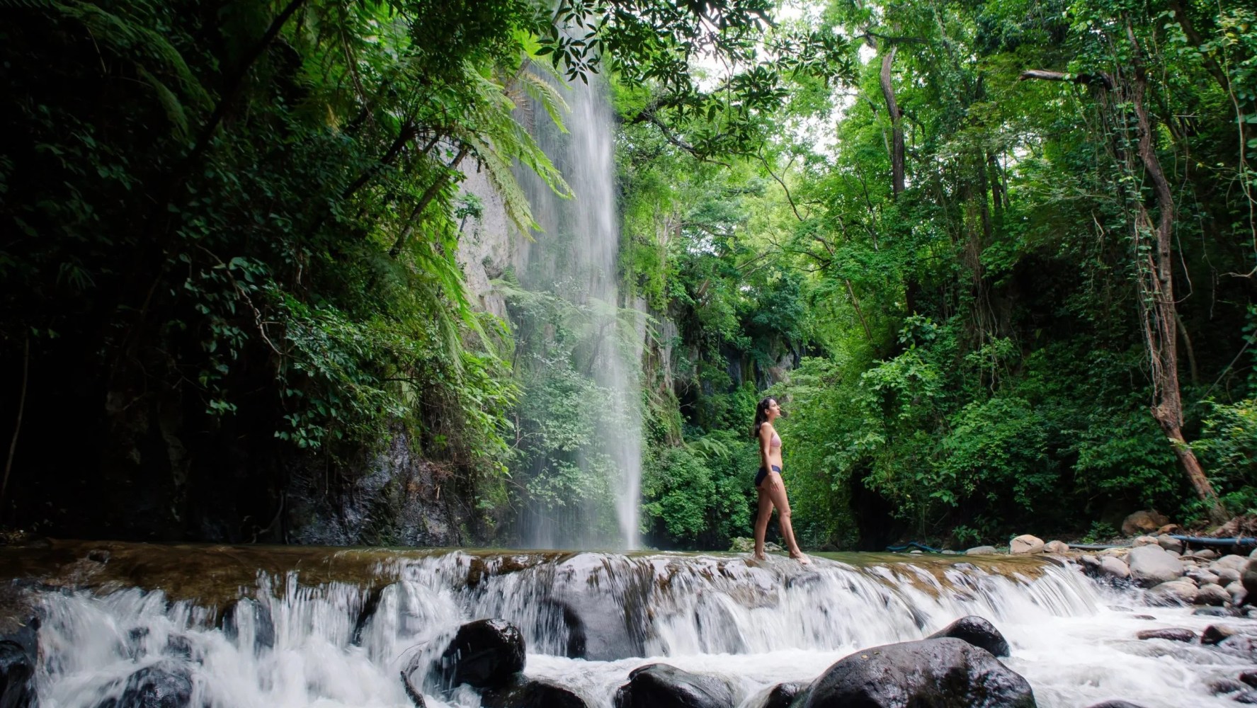 Person in swimwear stands near a waterfall in a lush forest.