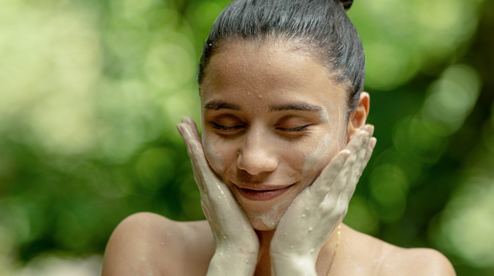 Woman with mud mask on face smiling and touching cheeks outdoors.