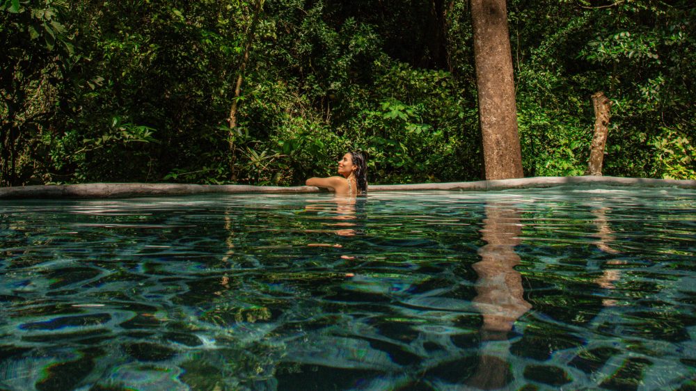 Person swimming in a clear pool surrounded by lush green trees.