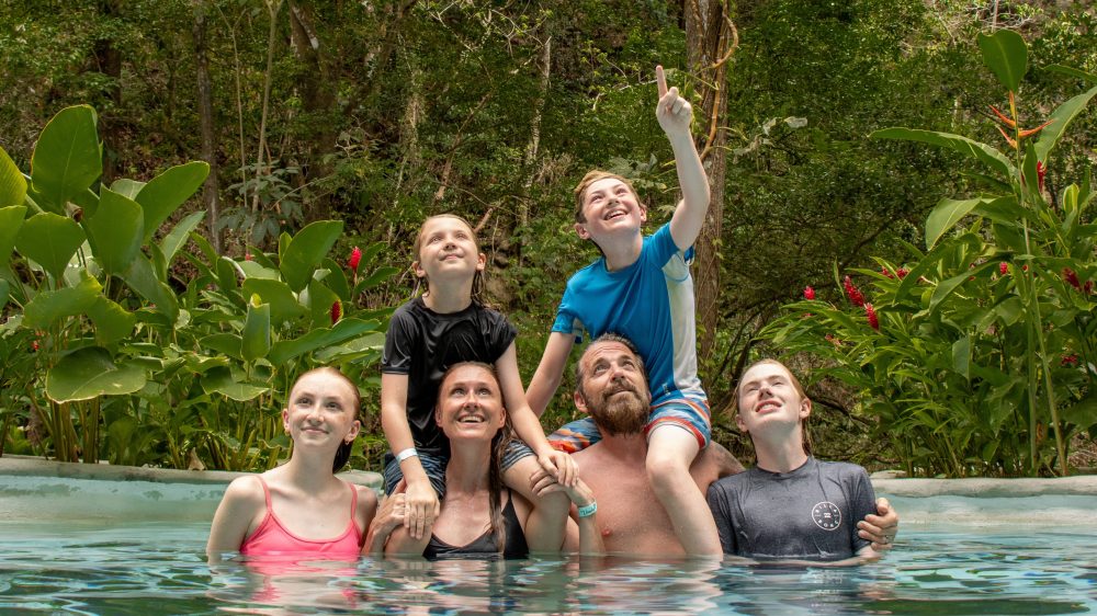 Family of six in a pool surrounded by lush greenery, two children on parents' shoulders, smiling.