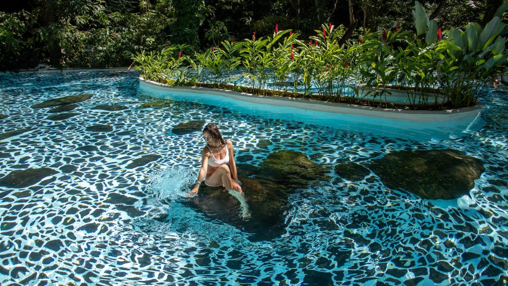 Woman in white bikini sits on a rock in a patterned pool with lush plants around.