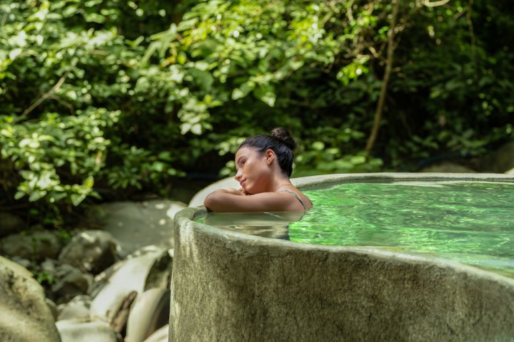 Person relaxing in a natural stone pool surrounded by lush greenery.